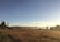A vast open prairie landscape with golden brown grasses under a clear blue sky. In the distance, mountains are visible with some mist hanging above them. On the left, a small wooden cabin is nestled among a few tall pine trees. The image has a serene and peaceful quality.