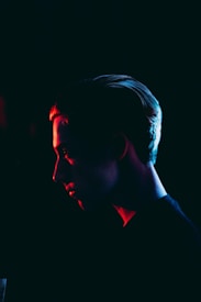 A person's side profile is highlighted in dramatic lighting, with a dark background. The face is partially illuminated by a red light, creating a mysterious and intense atmosphere.