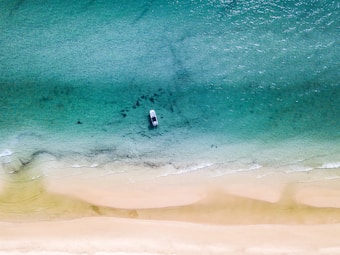 An aerial view of a boat on crystal clear turquoise water near a sandy beach. The gradient of the ocean transitions from deep teal to light aqua as it approaches the shore, with gentle waves visible. The boat is stationed close to the shore, highlighting the serenity and isolation of the setting.