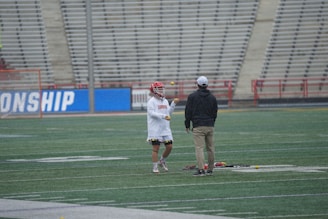 Two individuals are on a sports field. One person is wearing a red helmet and white athletic clothing, juggling balls. The other person is dressed in casual clothing with a cap and is observing. The field has a large empty seating area in the background.