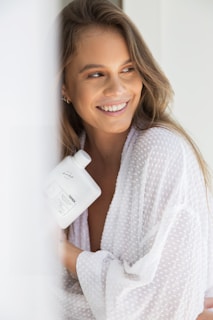 A smiling person with long hair is wearing a white textured robe while holding a white bottle with writing on it. The setting appears bright and soft, emphasizing a relaxed and cheerful ambiance.