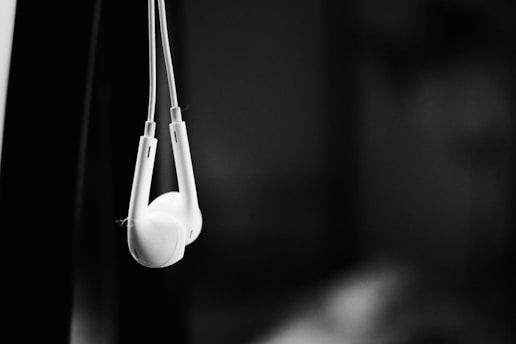 A pair of white earbuds hangs against a dark, blurred background, emphasizing the contrast between the bright white of the earbuds and the shadows behind them.