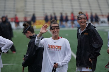 Several athletes in sports attire are on an outdoor sports field, interacting and smiling. One individual in the foreground waves with a friendly expression, while another stands beside her, also smiling. They wear protective goggles and gear associated with lacrosse. The background features indistinct figures in dark clothing against a backdrop of stadium seating.