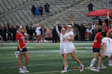 A group of female lacrosse players are on the field, with some celebrating and holding their sticks in the air. The players wear uniforms with distinct numbers and colors, red and white for one team and red and blue for the other. The background features stadium seating with some spectators.