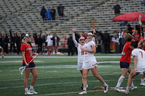 A group of female lacrosse players are on the field, with some celebrating and holding their sticks in the air. The players wear uniforms with distinct numbers and colors, red and white for one team and red and blue for the other. The background features stadium seating with some spectators.