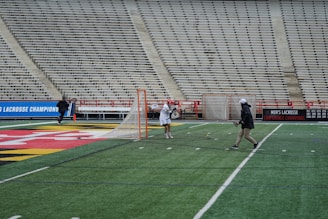 A lacrosse practice taking place on a field in a large, empty stadium. Two individuals are seen near the goal post, one wearing a red helmet and lacrosse gear and the other in casual attire with a hat. The field has lines and markings commonly found in sports arenas. There's a prominent banner on the stands highlighting men's lacrosse achievements.