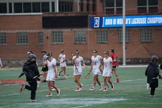 A group of female lacrosse players are walking on a field. They are wearing white uniforms with red accents. The building in the background has a banner that reads '2019 DI Women's Lacrosse Championship'. The field is artificial turf, and the weather appears overcast. A few other individuals are present, some in dark jackets, suggesting they might be coaches or officials.