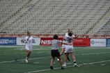 A group of female lacrosse players on a sports field with one player wearing a striped referee uniform in the foreground. Two players in white jerseys are hugging in celebration, while another player approaches with a smile, holding a lacrosse stick. The background shows empty stadium seating.