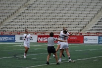 A group of female lacrosse players on a sports field with one player wearing a striped referee uniform in the foreground. Two players in white jerseys are hugging in celebration, while another player approaches with a smile, holding a lacrosse stick. The background shows empty stadium seating.
