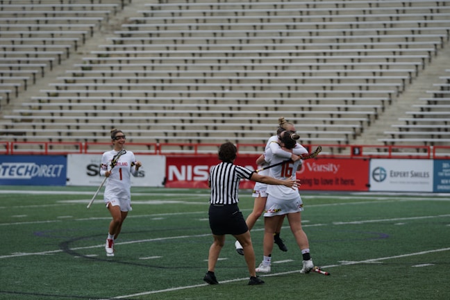 A group of female lacrosse players on a sports field with one player wearing a striped referee uniform in the foreground. Two players in white jerseys are hugging in celebration, while another player approaches with a smile, holding a lacrosse stick. The background shows empty stadium seating.