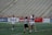 A group of female lacrosse players on a sports field with one player wearing a striped referee uniform in the foreground. Two players in white jerseys are hugging in celebration, while another player approaches with a smile, holding a lacrosse stick. The background shows empty stadium seating.