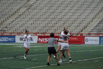 A group of female lacrosse players on a sports field with one player wearing a striped referee uniform in the foreground. Two players in white jerseys are hugging in celebration, while another player approaches with a smile, holding a lacrosse stick. The background shows empty stadium seating.