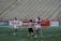 A group of female lacrosse players on a sports field with one player wearing a striped referee uniform in the foreground. Two players in white jerseys are hugging in celebration, while another player approaches with a smile, holding a lacrosse stick. The background shows empty stadium seating.