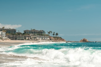 Waves crash onto a sandy beach with several modern beachfront houses in the background. Palm trees are interspersed among the buildings, and a rocky outcrop is visible further along the shoreline. The sky is clear with some clouds.