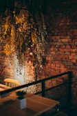 A rustic, warm interior with a brick wall enhanced by dried, hanging foliage. A wooden chair and table with a small potted plant suggest a cozy, informal setting.