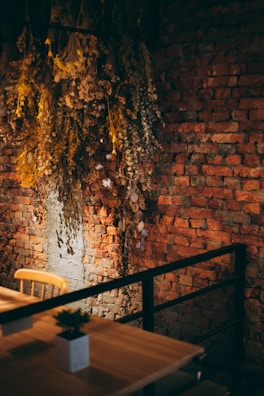 A rustic, warm interior with a brick wall enhanced by dried, hanging foliage. A wooden chair and table with a small potted plant suggest a cozy, informal setting.