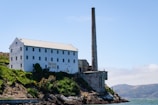 An industrial building with a tall chimney sits on a rocky coastline covered in lush greenery. The side of the building has graffiti, including the words 'Indian Land'. In the background, there are distant hills and a clear blue sky.