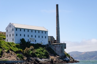 An industrial building with a tall chimney sits on a rocky coastline covered in lush greenery. The side of the building has graffiti, including the words 'Indian Land'. In the background, there are distant hills and a clear blue sky.