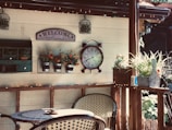 A cozy patio setting with wicker chairs and a small table. A wall features decorative items including a 'Welcome Friends' sign, a large vintage-style clock, and pots of vibrant flowers. There is a shelf with small plants and a rustic mailbox. Warm light from string lights creates a welcoming atmosphere.
