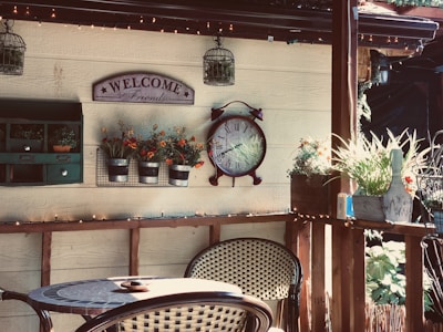A cozy patio setting with wicker chairs and a small table. A wall features decorative items including a 'Welcome Friends' sign, a large vintage-style clock, and pots of vibrant flowers. There is a shelf with small plants and a rustic mailbox. Warm light from string lights creates a welcoming atmosphere.