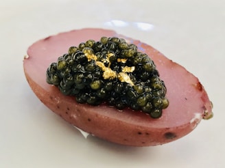 A piece of food featuring a smooth, oval slice of pinkish-purple potato topped with a rich layer of shiny black caviar and small flecks of gold leaf.
