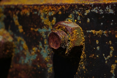 A close-up view of a rusty metal surface with patches of yellow and blue corrosion. The focus is on a large bolt, deeply corroded, with vibrant textures created by the oxidization process.