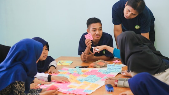 A group of people sitting around a table engaged in a discussion, with colorful sticky notes spread out in front of them. One person is pointing at a note, while others listen attentively. The setting appears to be a collaborative environment, possibly a workshop or brainstorming session.