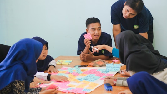 A group of people sitting around a table engaged in a discussion, with colorful sticky notes spread out in front of them. One person is pointing at a note, while others listen attentively. The setting appears to be a collaborative environment, possibly a workshop or brainstorming session.
