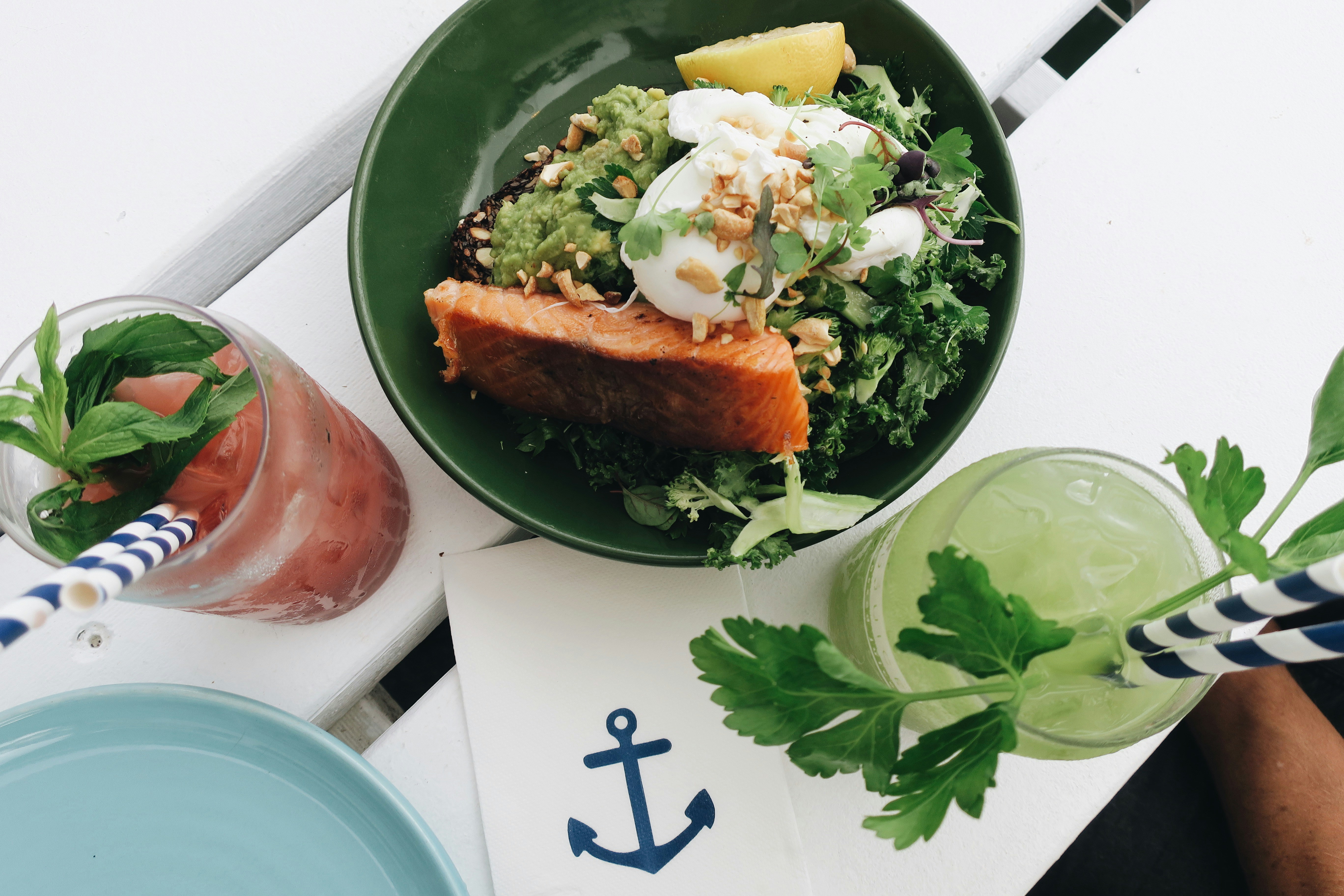 Top-down food photograph of a kale salad with a poached egg and nuts in a sea-green bowl, with citrus drinks on a white surface.