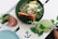 A green bowl filled with fresh salad topped with a piece of salmon, a dollop of cream, and garnished with herbs and nuts. Beside it, there are two beverages: one red with mint and a blue and white striped straw, and another green with a similar straw. A decorative napkin with a blue anchor design sits under the bowl.