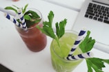 Two refreshing beverages are placed side by side on a white surface. One drink is a reddish-brown color, garnished with fresh mint leaves, and the other is a light green color, garnished with parsley. Both drinks have blue and white striped straws. In the background, the edge of a laptop is visible.