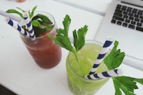 Two refreshing beverages are placed side by side on a white surface. One drink is a reddish-brown color, garnished with fresh mint leaves, and the other is a light green color, garnished with parsley. Both drinks have blue and white striped straws. In the background, the edge of a laptop is visible.