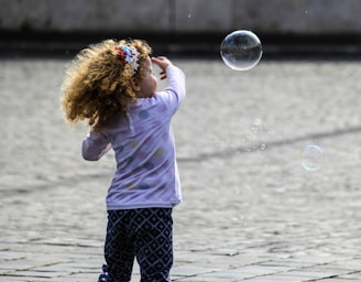 A young child with curly hair and a colorful headband is playfully reaching towards floating bubbles in an open space. The ground is paved with large stones, and the child is wearing a polka-dot long-sleeve shirt and patterned pants.