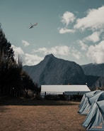 A scenic landscape with a helicopter flying over a mountain range in the background. The foreground features several pitched tents on a grassy area, with a building that has a metal roof situated nearby. Trees are visible on the left side, adding a sense of natural serenity to the scene.