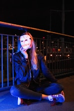 A person seated cross-legged on the ground holding a white mask with a sinister smile in front of their face. The scene is dimly lit with blue and orange lights, and the background features metal railings and what appears to be city lights or reflections.