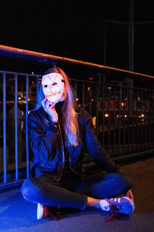 A person seated cross-legged on the ground holding a white mask with a sinister smile in front of their face. The scene is dimly lit with blue and orange lights, and the background features metal railings and what appears to be city lights or reflections.
