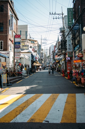 A bustling urban street filled with various shops and signs, lined with red brick buildings and crisscrossed with overhead utility wires. Pedestrians walk along the street while colorful signs in different languages advertise eateries and stores along the sidewalk. The atmosphere conveys a busy, lively commercial district.