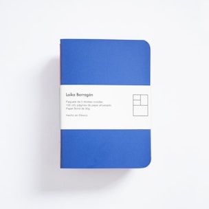 A blue notebook with rounded corners is wrapped in a white paper band. The band contains text in Spanish describing the product as a package of five sewn notebooks with 100 pages each, made with 90g bond paper, manufactured in Mexico. The design includes a minimalist, geometric logo.