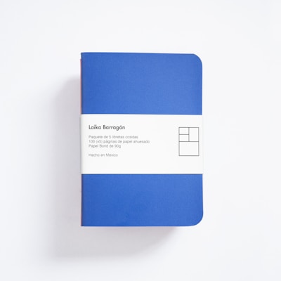 A blue notebook with rounded corners is wrapped in a white paper band. The band contains text in Spanish describing the product as a package of five sewn notebooks with 100 pages each, made with 90g bond paper, manufactured in Mexico. The design includes a minimalist, geometric logo.