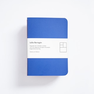 A blue notebook with rounded corners is wrapped in a white paper band. The band contains text in Spanish describing the product as a package of five sewn notebooks with 100 pages each, made with 90g bond paper, manufactured in Mexico. The design includes a minimalist, geometric logo.