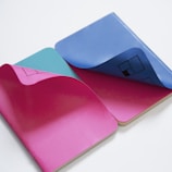 Two colorful notebooks with open pages showing vibrant pink, blue, and teal colors. The pages are curling slightly, revealing geometric patterns and designs printed on the inside.