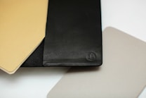 A collection of flat, rectangular objects overlapping each other, including a black leather item with a logo embossed on it, as well as beige and grey textured pieces.
