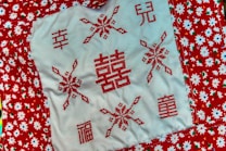 A piece of cloth features a central white section adorned with red cross-stitch embroidery depicting various Chinese characters and snowflake-like patterns. The surrounding fabric is red with a white floral pattern, featuring green leaves and blue centers.