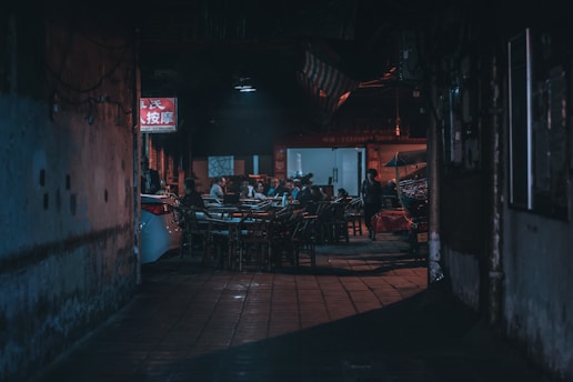 A dimly lit alley with an outdoor dining area featuring several people sitting around tables. The scene includes a narrow, urban setting with a neon sign, a parked car, and various chairs. The environment appears cozy yet slightly mysterious due to the lighting.