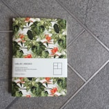 A notebook with a tropical plant design cover, featuring large green leaves and vibrant orange flowers. The notebook's cover is partially wrapped with a white paper band that contains text and a simplistic graphic. The notebook is placed on a textured, gray tiled surface.