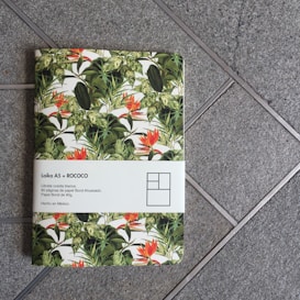 A notebook with a tropical plant design cover, featuring large green leaves and vibrant orange flowers. The notebook's cover is partially wrapped with a white paper band that contains text and a simplistic graphic. The notebook is placed on a textured, gray tiled surface.