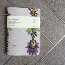 A notebook with a colorful tropical design sits on a tiled floor. The cover features illustrations of vibrant leaves, a bird, and a patterned background. A white band around the notebook displays text in Spanish, mentioning 'Laika A5 + ROCOCO' and details about the notebook's paper.