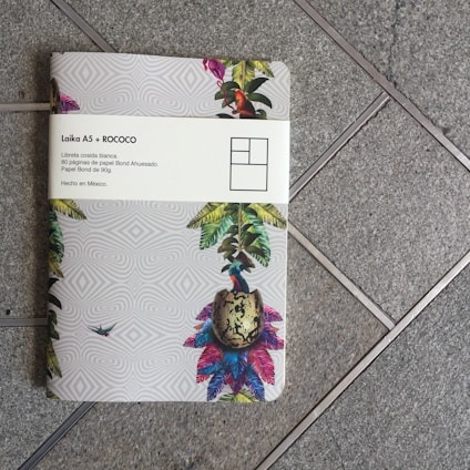 A notebook with a colorful tropical design sits on a tiled floor. The cover features illustrations of vibrant leaves, a bird, and a patterned background. A white band around the notebook displays text in Spanish, mentioning 'Laika A5 + ROCOCO' and details about the notebook's paper.