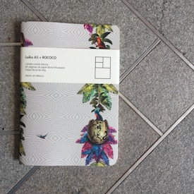 A notebook with a colorful tropical design sits on a tiled floor. The cover features illustrations of vibrant leaves, a bird, and a patterned background. A white band around the notebook displays text in Spanish, mentioning 'Laika A5 + ROCOCO' and details about the notebook's paper.