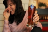 A person wearing a striped shirt is laughing while holding a tall glass of beer. The beer glass has a label with the text 'HB MÜNCHEN' and a crown symbol. The background appears blurred, with hints of warm lighting and indoor decorations.
